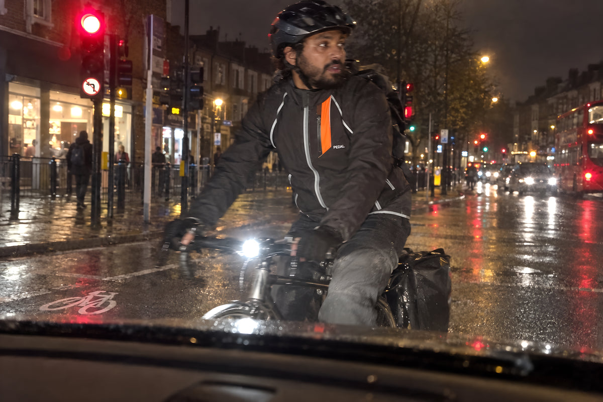 A cyclist in the black PEDAL Jacket with its orange tab crossing in front of a stopped car at an intersection at night, seen from the driver's seat through the windshield. The rider is close, large in the frame and clearly lit by the car's headlights: the jacket's reflective strips and orange tab lit up, the face readable, the PEDAL front light bright. Traffic lights and shop signs behind.