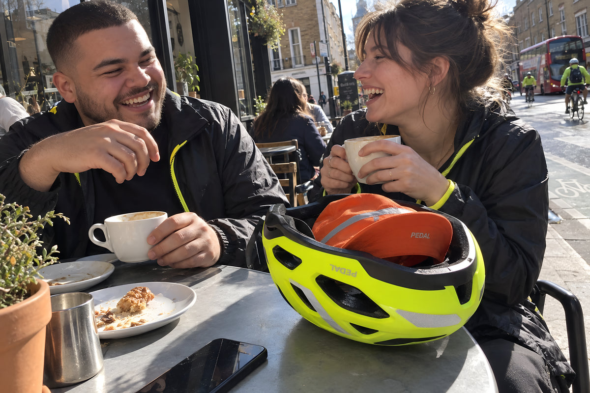 Two riders at an outside cafe table, the PEDAL Helmet upside down on the table with the orange PEDAL Cap stuffed in it, both mid-laugh over coffee.