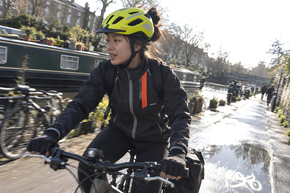A woman riding a canal towpath in the fluorescent yellow PEDAL Helmet and black Jacket, a narrowboat and moored bikes going past, dodging a puddle.