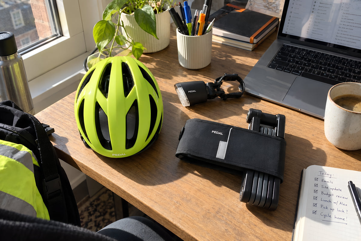 The PEDAL Commute Kit tipped out on an office desk beside a laptop, helmet, lock and lights among the pens and a coffee cup.