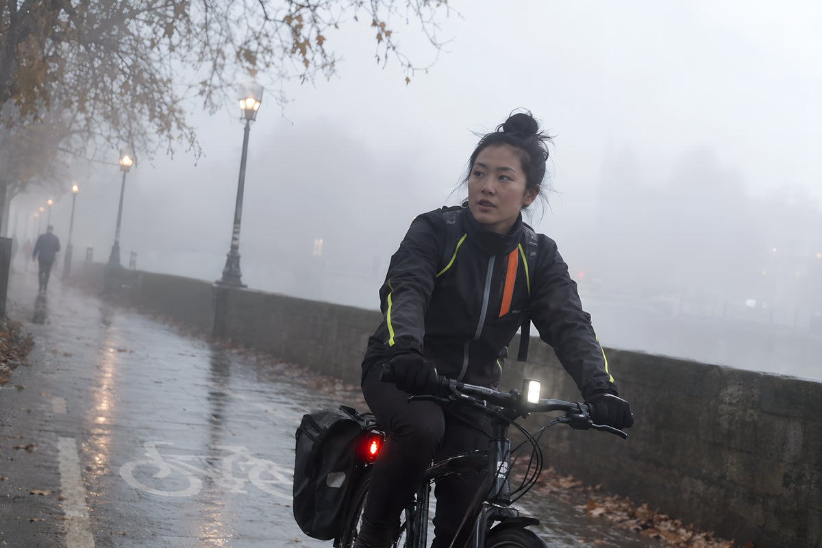 A cyclist emerging out of thick morning fog on a park path, only the fluorescent trim and the rear light clearly readable at distance.