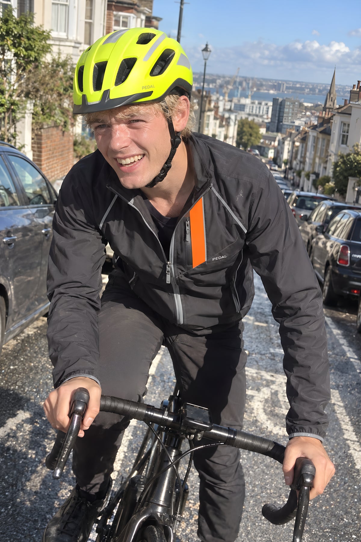 A man out of the saddle grinding up a steep street of row houses in the PEDAL Jacket and Helmet, jacket open at the neck, parked cars either side.