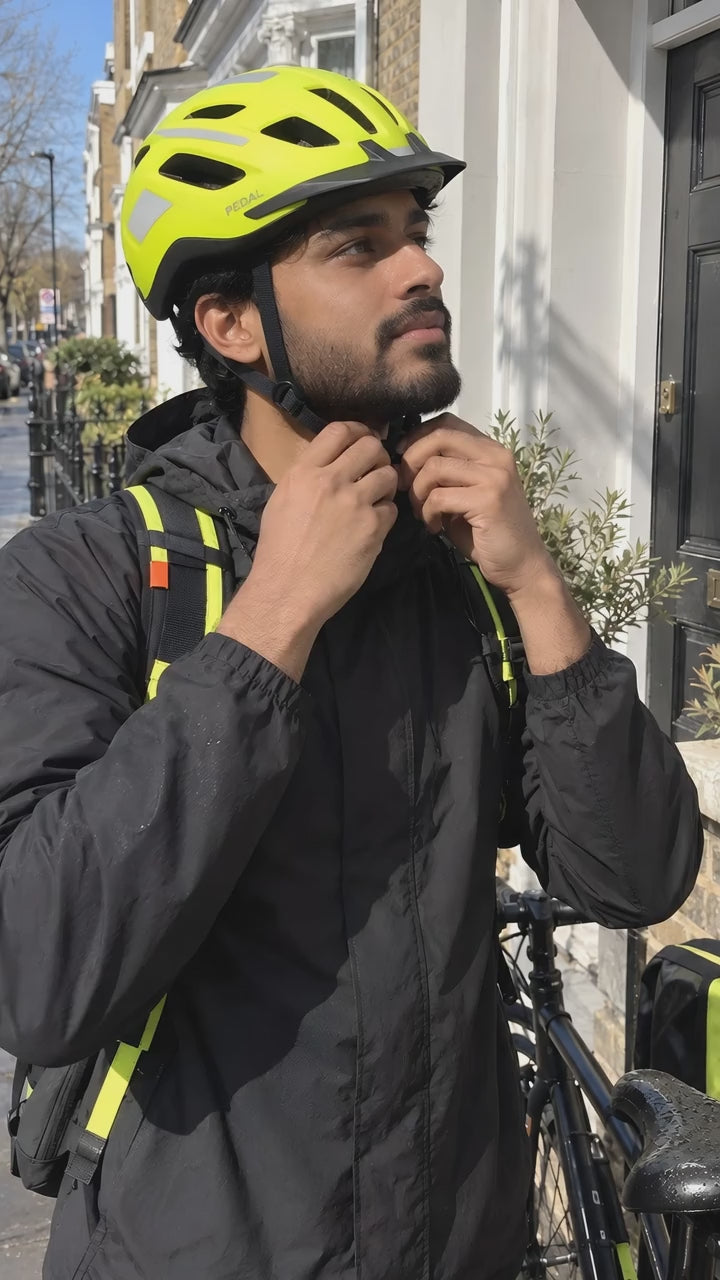 A man doing up the chin strap of the yellow PEDAL Helmet outside his front door